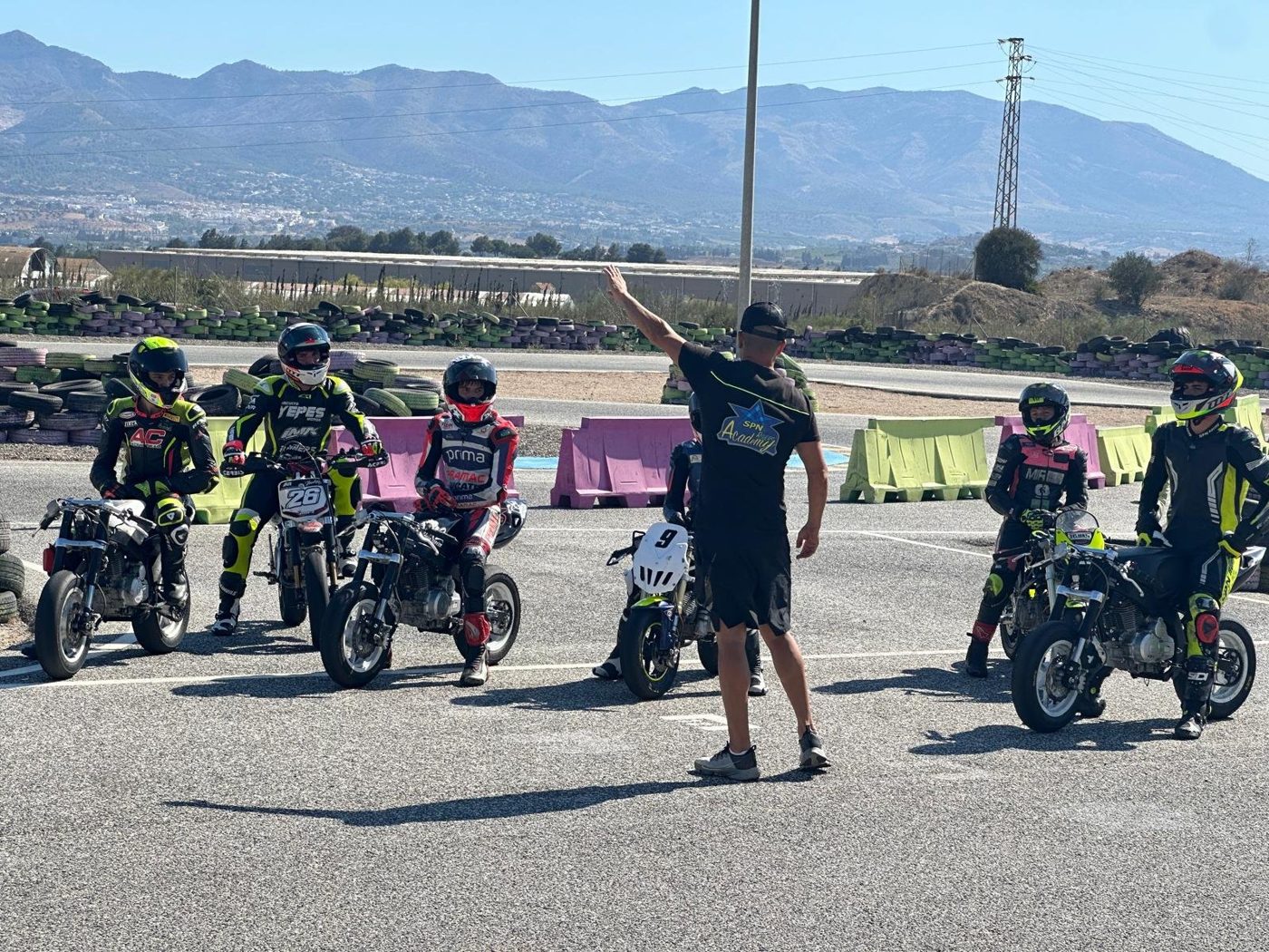 Coach Nico Ferreira, in an SPN Academy shirt, briefs a line of riders on small bikes before a session, with mountains behind the track.