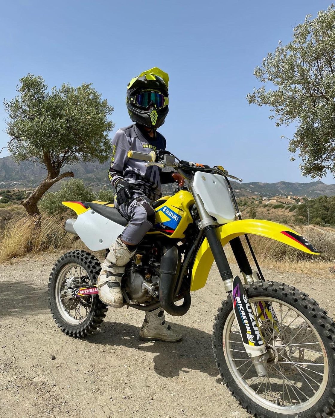 Josh in a yellow helmet and grey riding kit, seated on a yellow Suzuki dirt bike on a dirt track with olive trees and mountains behind.