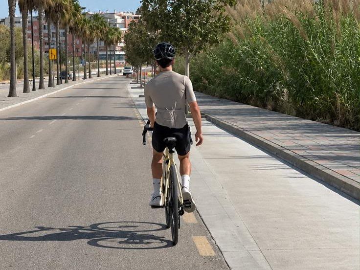 Josh from behind on a road bike, riding down a palm-lined road in a helmet and cycling kit.