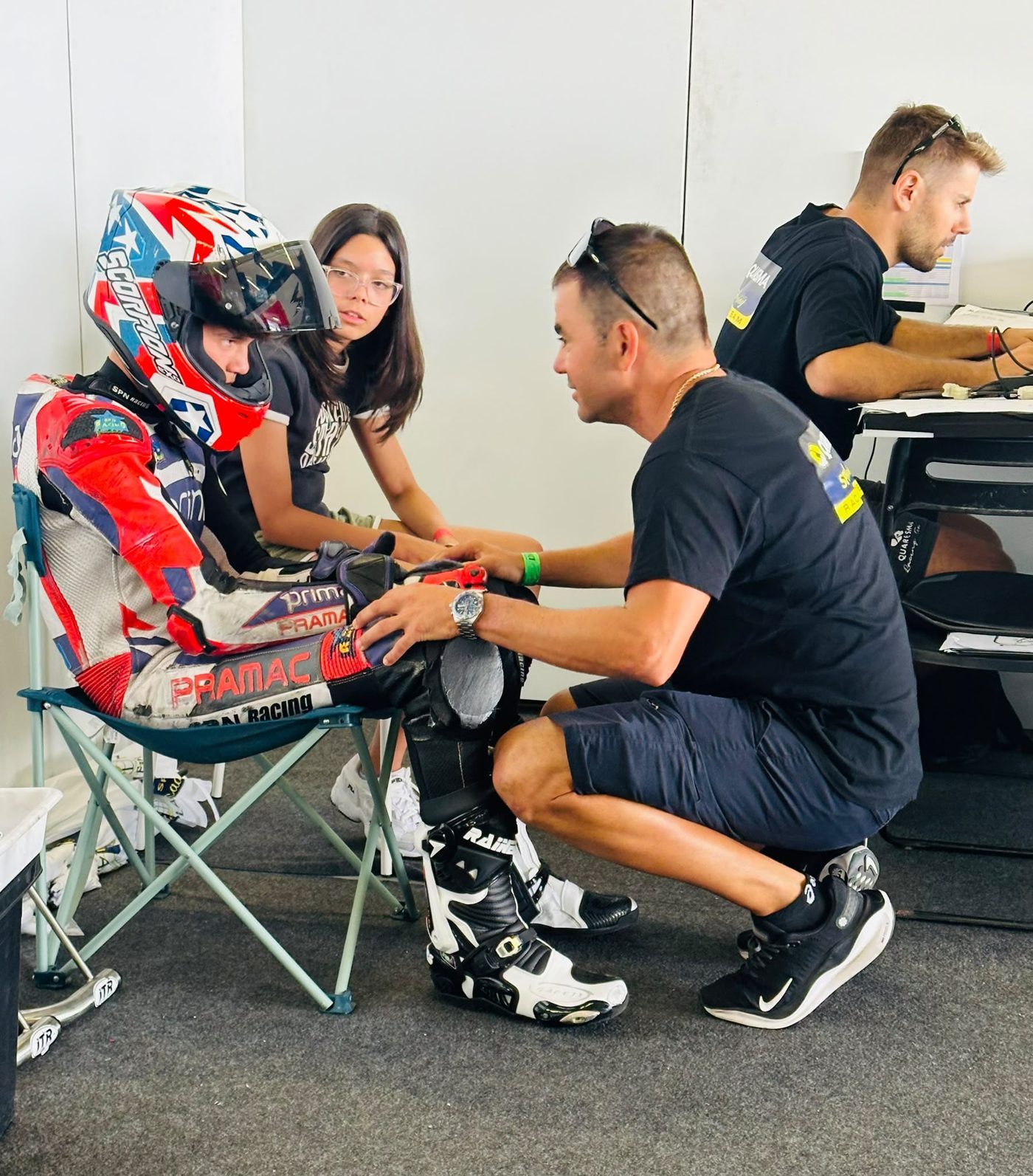 Josh in his race leathers and helmet, seated in the paddock while his coach crouches beside him with a hand on his knee.