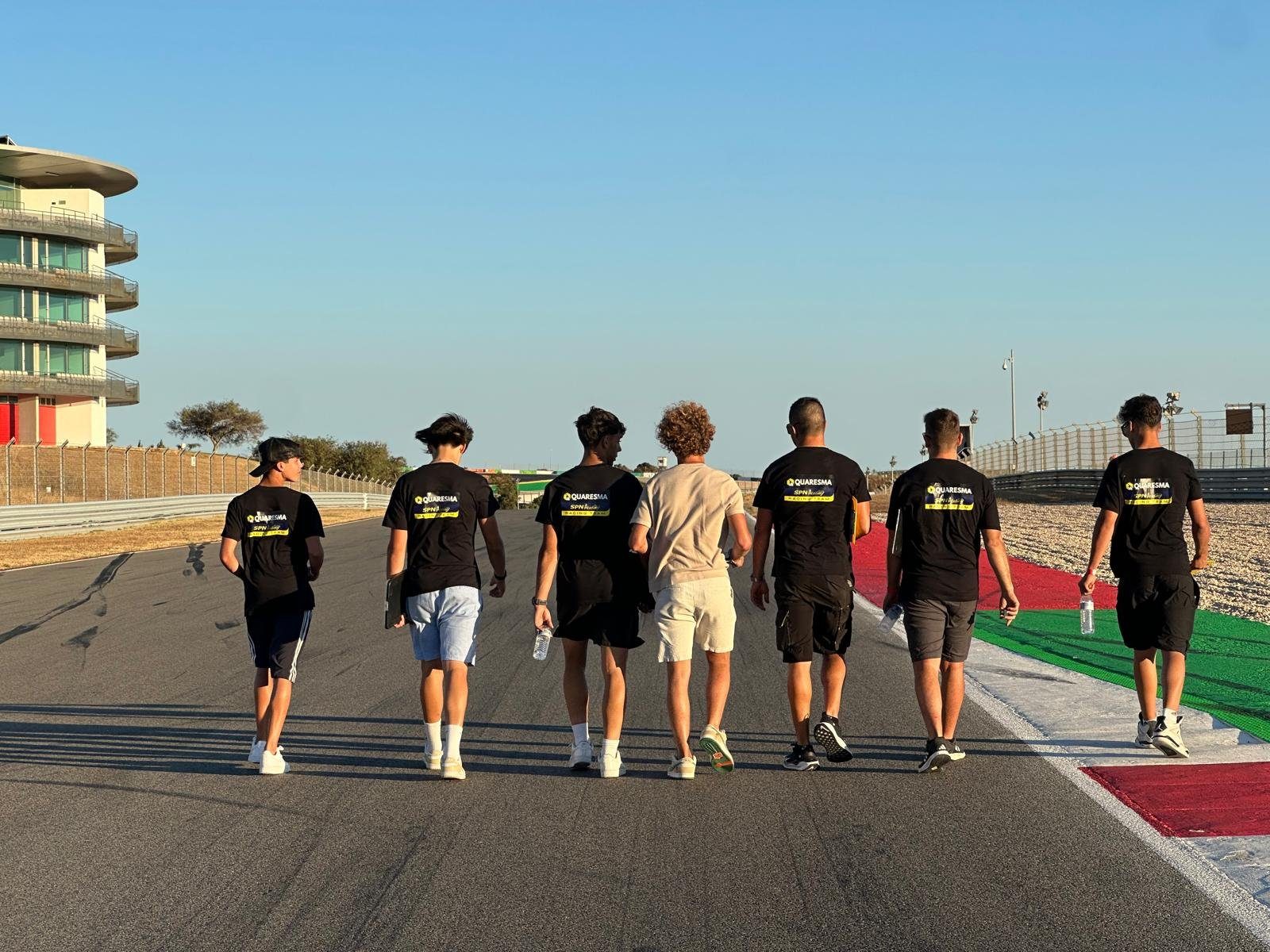 Josh and the Quaresma SPN Academy Racing Team, in matching black team shirts, walk away from the camera along the empty circuit at Portimão, with the tower building on the left.