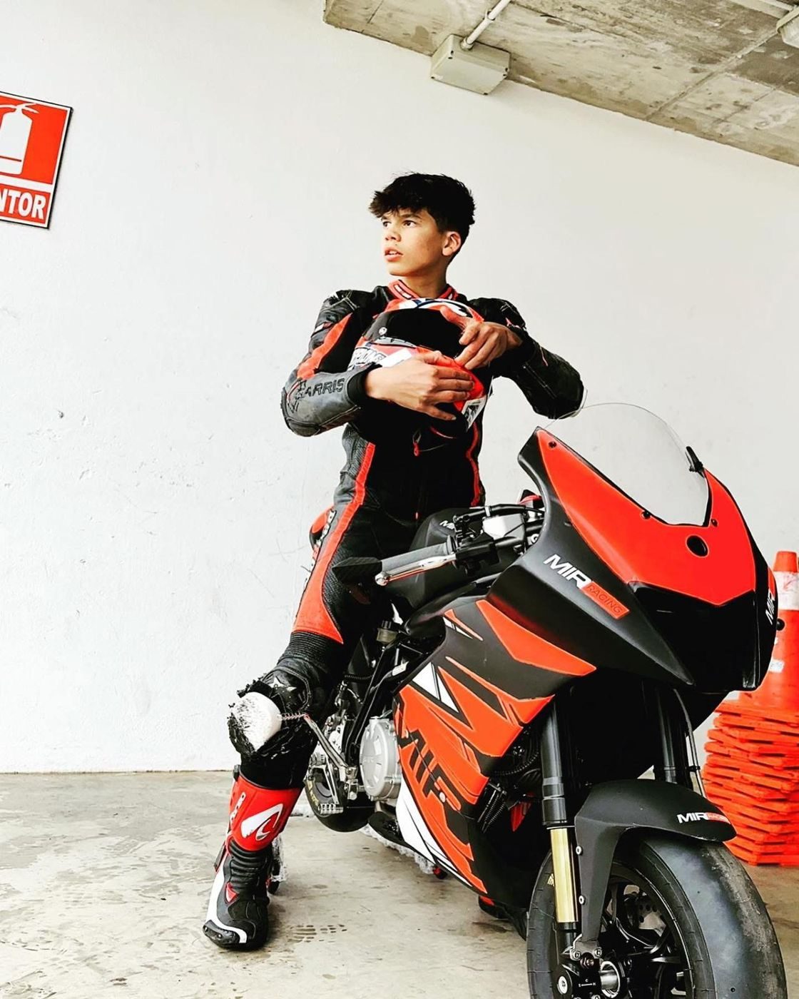 Josh in red and black leathers holding his helmet, leaning on an orange and black MIR bike in the garage.