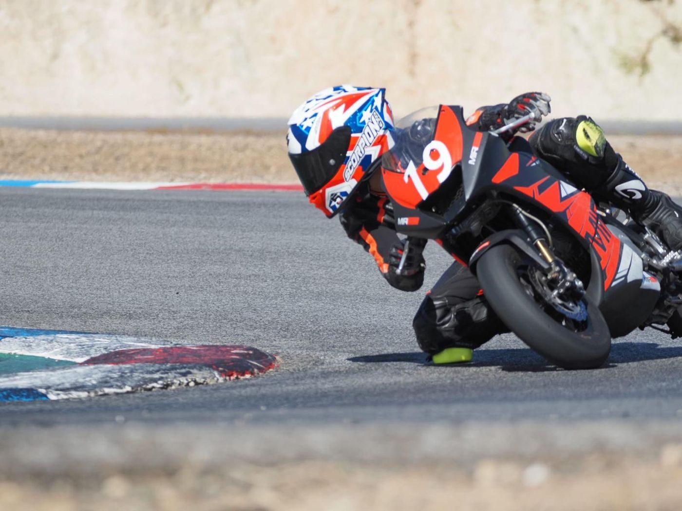 Josh at full lean on the orange and black MIR bike, number 19, his knee near the kerb.