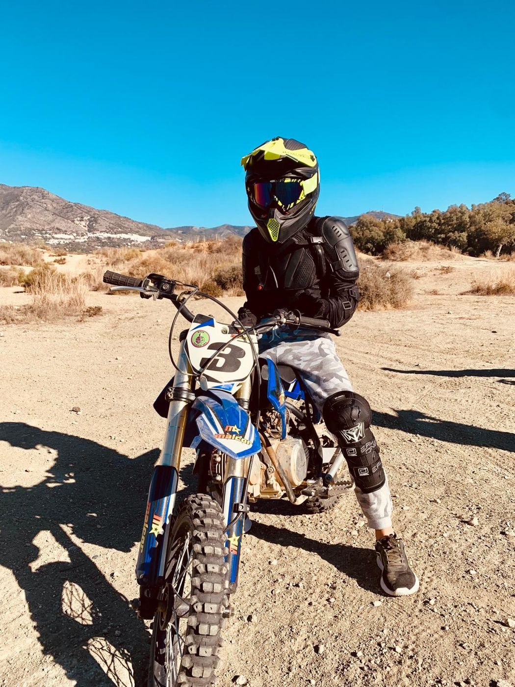 Josh in a yellow helmet, goggles and body armour, seated on a blue dirt bike with the number 3, on dry ground with mountains behind.