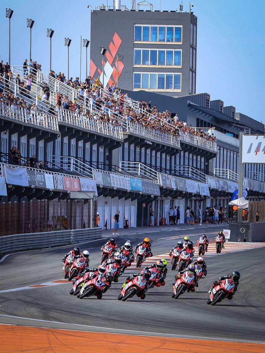 A full PreMoto3 field on red bikes leaning into the first corner after the start, with packed grandstands above the pit garages.
