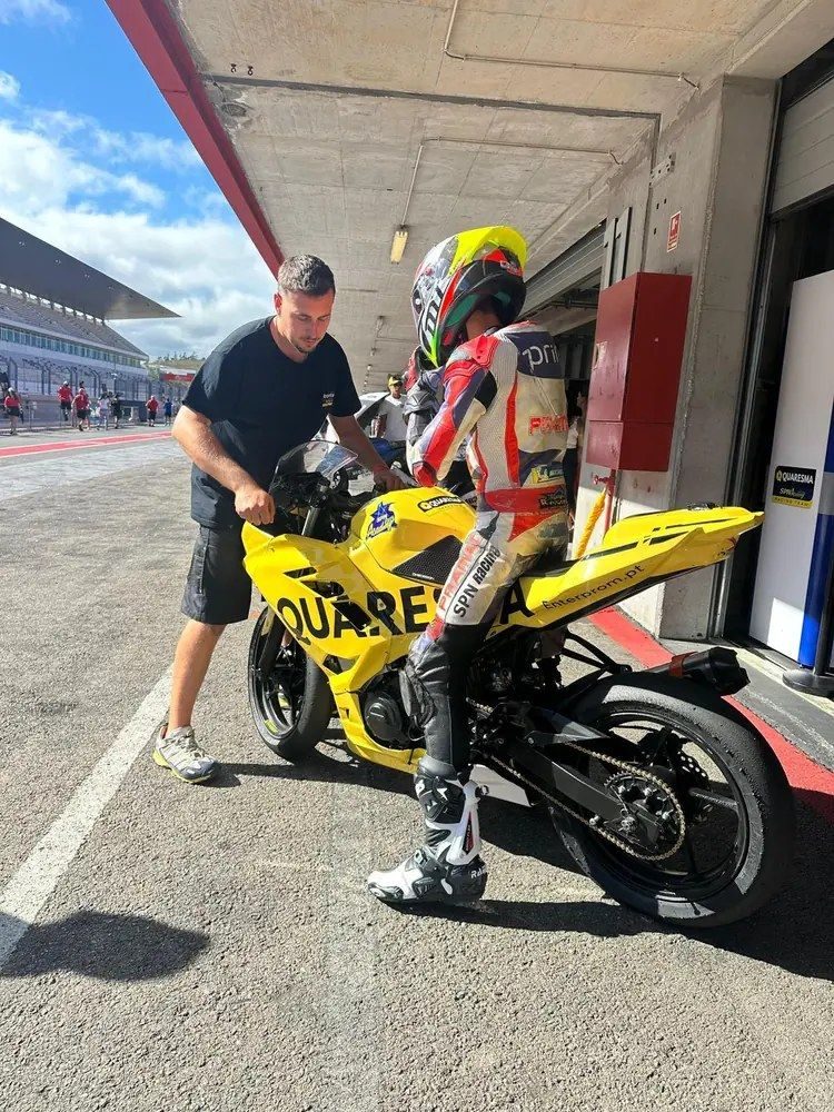 Josh in his leathers in the pit garage beside the yellow race bike, with a mechanic beside him.