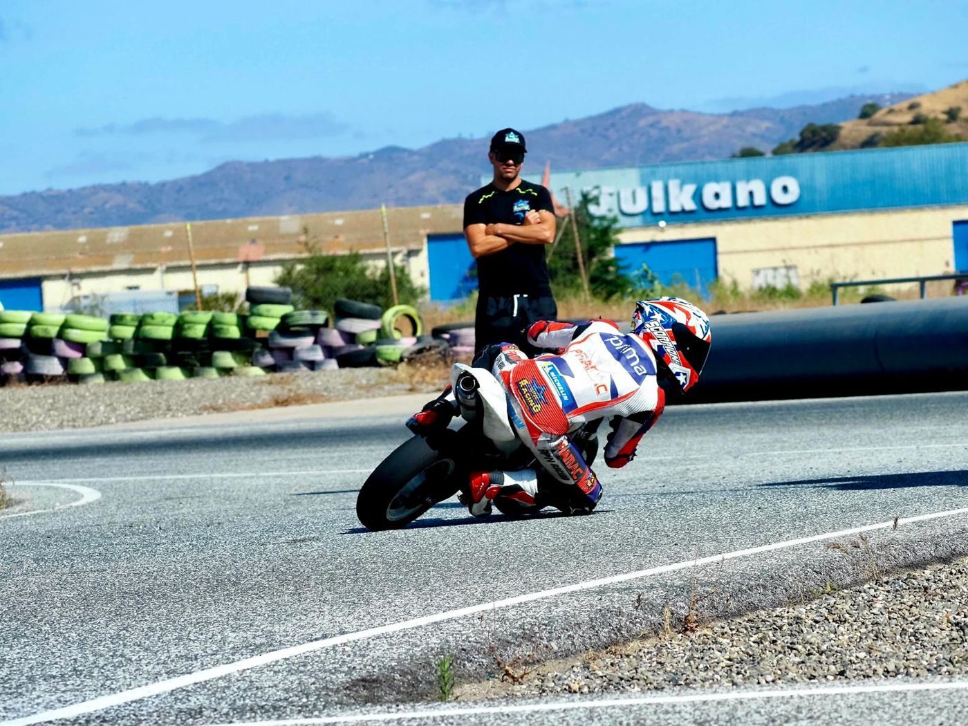 Josh in white, red and blue leathers leaning a small bike into a tight corner while a coach watches with arms crossed.