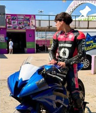 Josh standing beside a blue bike in the paddock in his MIR Racing leathers.