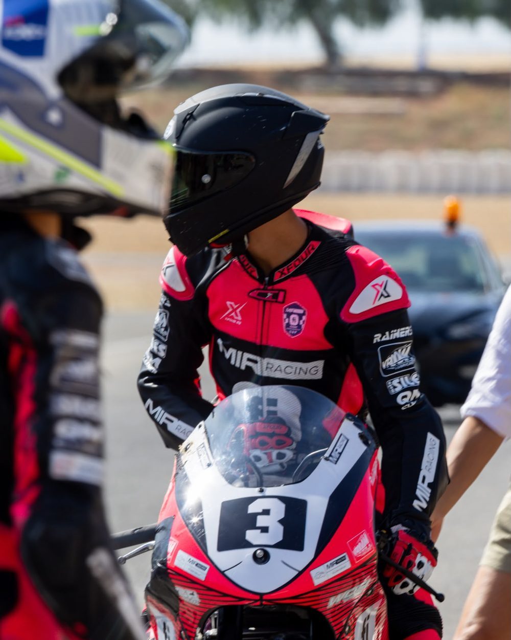 Josh in the paddock in his pink and black MIR Racing leathers and black helmet, looking back over his shoulder from the red bike.